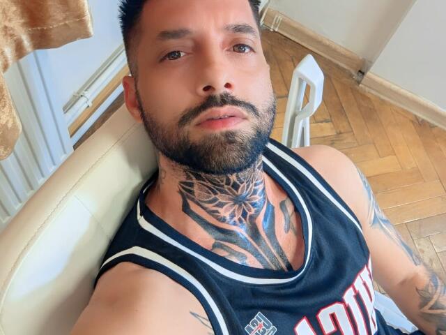TommyDum, con una maglietta da basket bianco e nero, tatuaggi sul collo e sul petto, capelli scuri corti e barba, seduto su una sedia bianca, pavimento in legno..