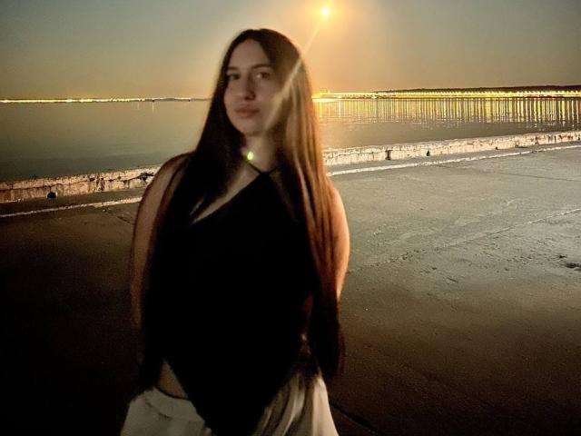Gearldine Gyles, con lunghi capelli castani, indossa una canotta nera e pantaloni bianchi, ed è in piedi su una spiaggia di sabbia scura al tramonto, con il sole che le illumina la schiena..