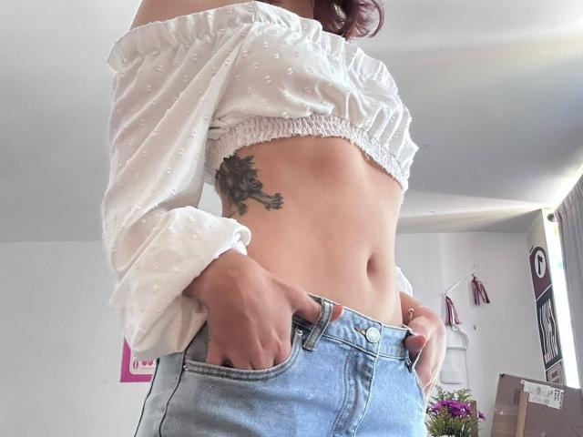 SpencerLe with a white off-shoulder crop top, exposing a toned midriff and tattoo on the right side, wearing light blue jeans with hands in pockets.