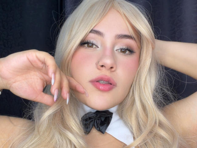 LorensDouce, con cabello rubio, labios rosados y uñas largas, vestida con una pajarita negra y un cuello blanco, mirando seductoramente a la cámara, con una mano tocándose la mejilla..