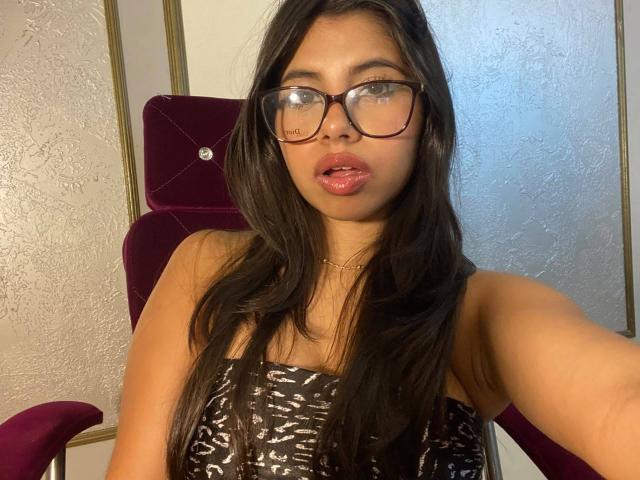 Celeste Morel, avec ses longs cheveux noirs, ses lunettes et sa robe noire à motifs, a des lèvres roses et pulpeuses et porte un collier en or, assise sur une chaise violette..
