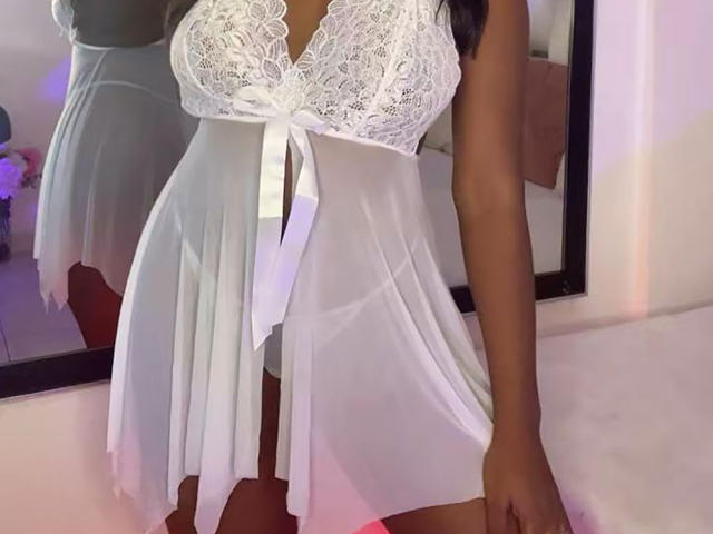 VenusWilly portant une nuisette blanche en dentelle et en tissu transparent, avec un ruban, ses seins étant partiellement visibles à travers le tissu délicat, debout devant un miroir..