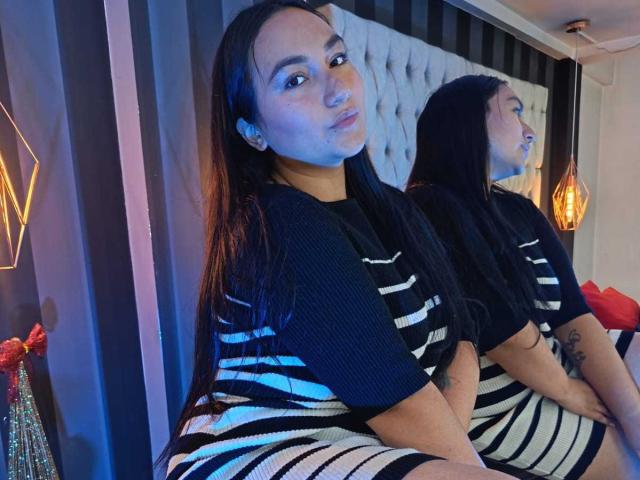 JeniferMorat with long black hair, wearing a black and white striped dress, sitting on a bed, reflection in mirror showing her pouty face and tattoo on right arm.