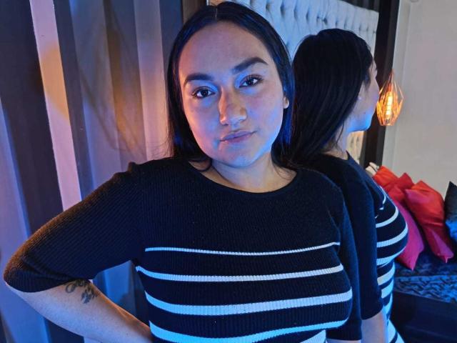 JeniferMorat with straight black hair, wearing a black and white striped sweater, standing in front of a mirror, showing her reflection and a tattoo on her arm.