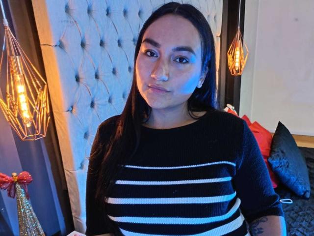 JeniferMorat with long black hair, wearing a black and white striped sweater, standing in a room with blue lighting, looking at the camera with a slight smile.