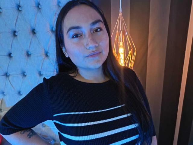 JeniferMorat with long dark hair, wearing a black striped shirt, standing with hands on hips, has a tattoo on her right arm, and a warm light behind her.