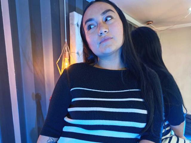 JeniferMorat has long black hair, a serious expression, and is wearing a black and white striped sweater. Her reflection is in the mirror behind her.