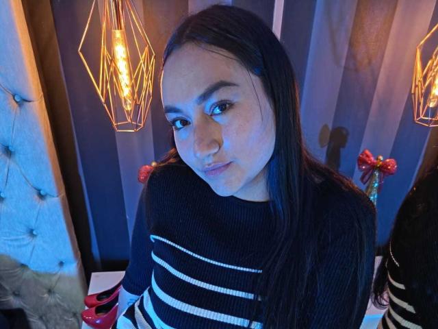 JeniferMorat with long dark hair, wearing a black and white striped sweater, looking down with a slight smile, under geometric hanging lights. Her ass is not visible.