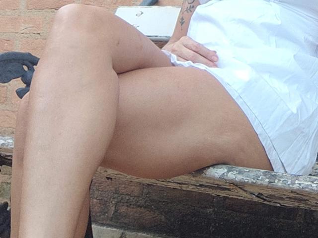 De witte shorts van RainhaDeusa, haar gebruinde, gespierde benen gekruist, onthullen haar gladde, kale dijen. Ze zit op een ruw, houten oppervlak en er is een tatoeage zichtbaar op haar arm..