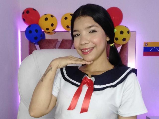 TaylorGil with black pigtails, smiling, wearing a white sailor uniform with a red ribbon, hand on chin, colorful balloons behind her, small tattoo on right wrist.