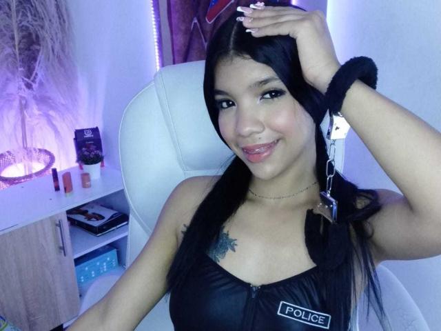 TaylorGil with black hair, small tattoo on chest, black police uniform, handcuffs on wrist, smiling, holding phone above head, sitting in white chair, blue light in background.