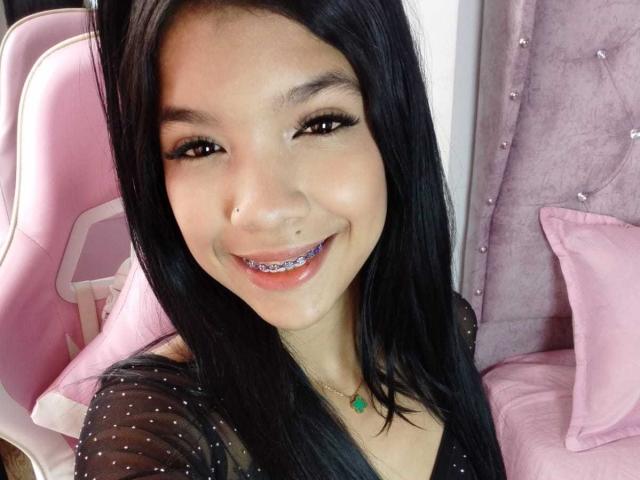 TaylorGil has black hair, brown eyes, and a nose ring. She's smiling with purple braces, wearing a sheer black top, and a green necklace, sitting in a pink chair.