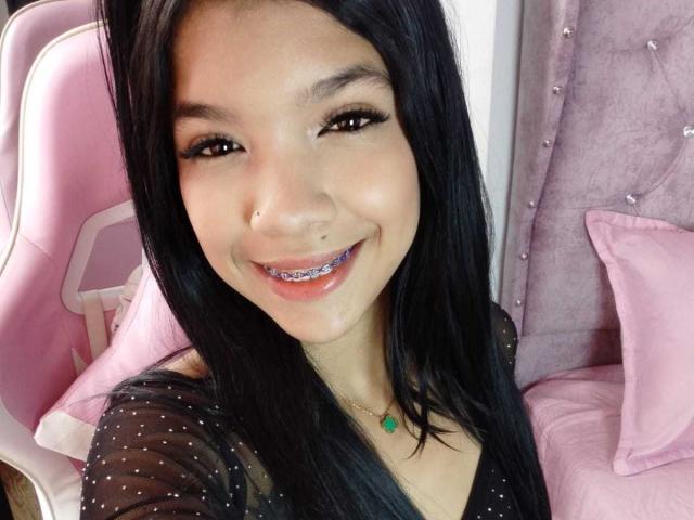 TaylorGil with black hair, smiling with metallic braces, wearing a sheer black top, and a green necklace, sitting in a pink chair, showing her nose piercing.