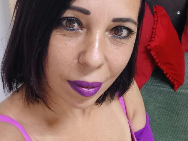 LauraSin has shiny purple lipstick, dark brown eyes with heavy eyeliner, black shoulder-length hair, and is wearing a purple top, looking up with a slight smile.
