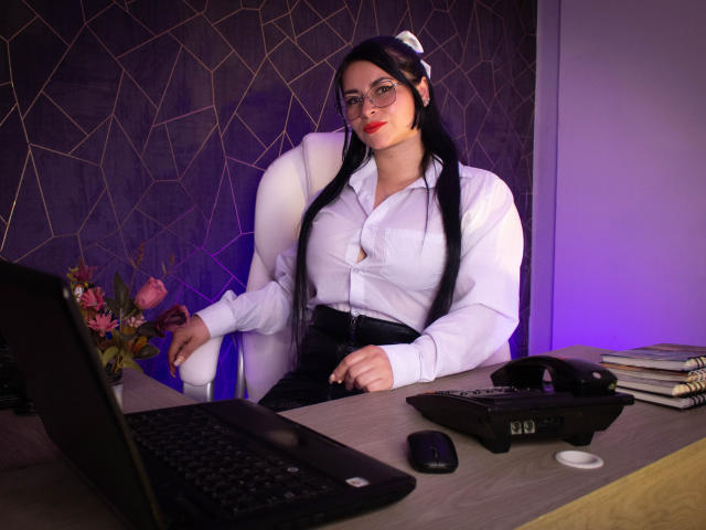 Nicole Memphis, mit langem, schwarzem Haar, Brille und rotem Lippenstift, sitzt in einem weißen Bürostuhl, trägt eine weiße Bluse und einen schwarzen Rock und befindet sich an einem Schreibtisch mit einem Laptop..