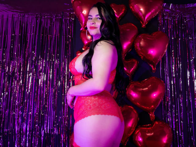 NicoleMemphis con un gran trasero, senos grandes, en lencería de encaje rojo, cabello negro largo, iluminación púrpura, de pie junto a globos rojos brillantes en forma de corazón, mirando a la cámara..