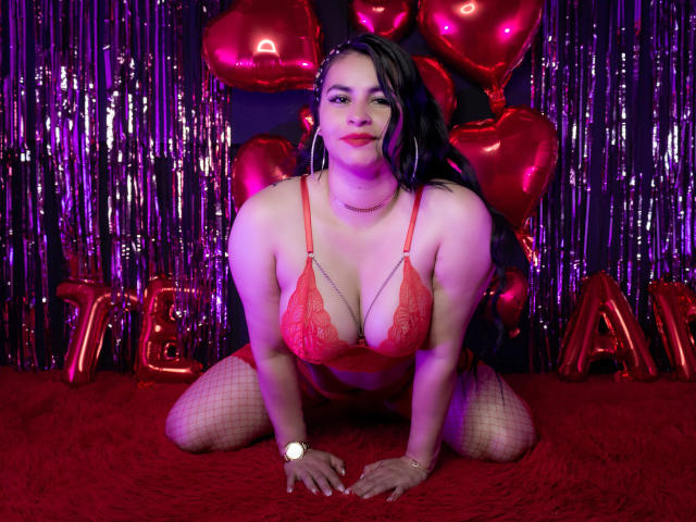 Nicole, kniend, trägt einen roten Spitzen-BH, Netzstrümpfe und große Ohrringe, mit großen, runden Brüsten und einem selbstbewussten, sexy Ausdruck, umgeben von roten Herzballons und metallischen Girlanden..
