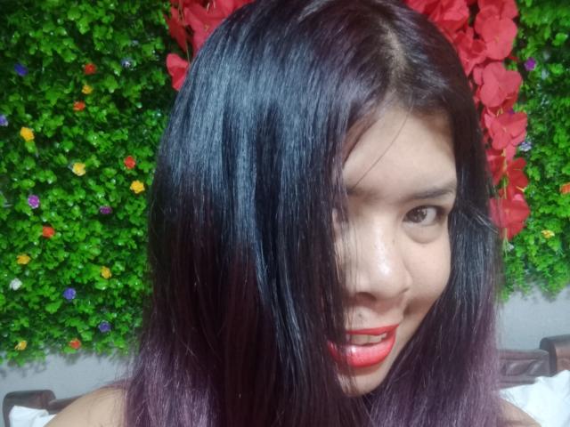 MariahSquirting69 with shiny black-purple hair, red lipstick, and a cute smile, looking at the camera, her tits slightly visible in the shot.