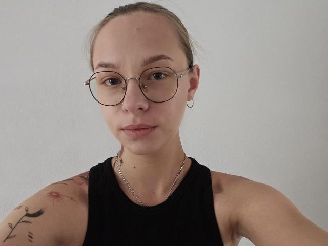 SteshaGlass with glasses, black tank top, tattoo on right shoulder, small nose ring, chain necklace, and subtle smile, looking at camera.