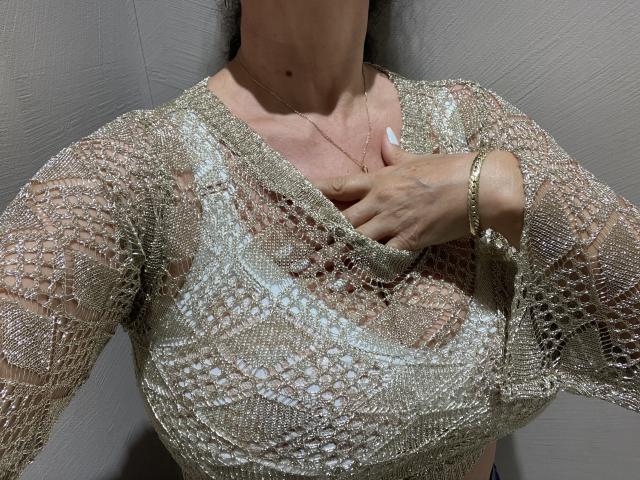 DeesseBleue's titty's are covered in a sheer, gold, lacey top, with her hand gently touching her chest. She's wearing a gold bracelet and a delicate necklace.