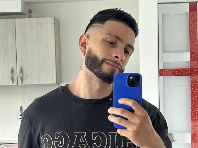 MarkBuffett, con una barba corta e scura, una camicia nera e un telefono blu, lo tiene in alto per scattare un selfie, mostrando un piccolo piercing sul lato sinistro della guancia..