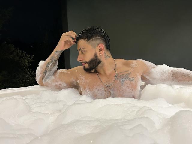 MarkBuffett, shirtless with soapy bubbles covering his chest, has a tattoo on his right shoulder, short black hair, and a beard, sitting in a bubbling hot tub at night.