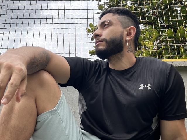 MarkBuffett, musculoso, con barba, camisa negra Under Armour, tatuajes, aretes, cabello corto y oscuro, pantalones cortos verdes, sentado con la rodilla levantada, mirando hacia un lado, una red detrás de él..