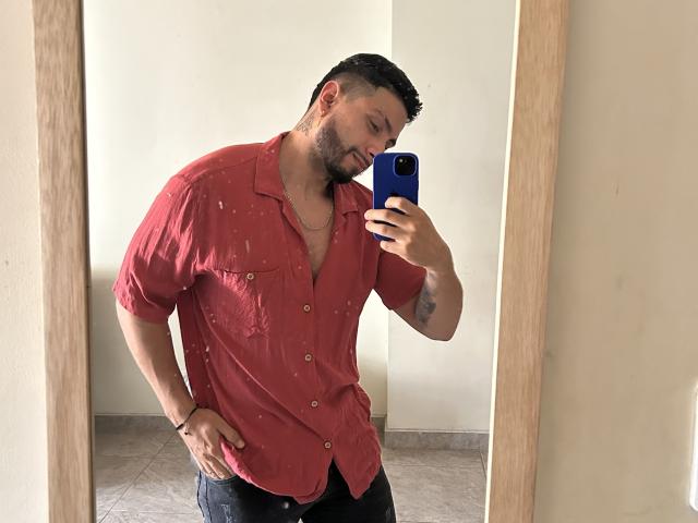MarkBuffett, muscular, bearded, wearing a red, button-up shirt, black pants, holding a blue phone, taking a mirror selfie, wood-framed mirror, tattoo on right forearm, casual, confident.