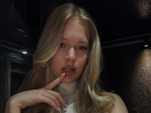 LillianaWhang has blonde hair, light skin, and is sucking her finger with her lips slightly parted. She's wearing a white turtleneck, and her finger is near her mouth.