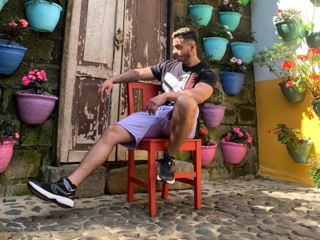 MarkBuffett, con tatuajes y barba, sentado en una silla roja, usando zapatillas negras y blancas, pantalones cortos morados y una camiseta negra, en un lugar iluminado por el sol, con adoquines y lleno de flores..