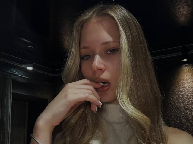 LillianaWhang with blonde hair, green eyes, and a white turtleneck, biting her finger, showing a small hint of her tits, in a dimly lit room.