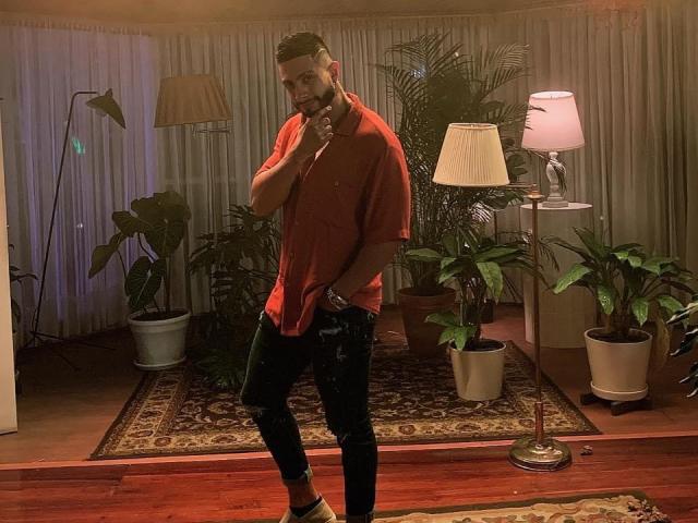 MarkBuffett stands in a room, wearing a red shirt, black pants, and white socks, with a beard, holding his chin, surrounded by plants and lamps, looking serious.