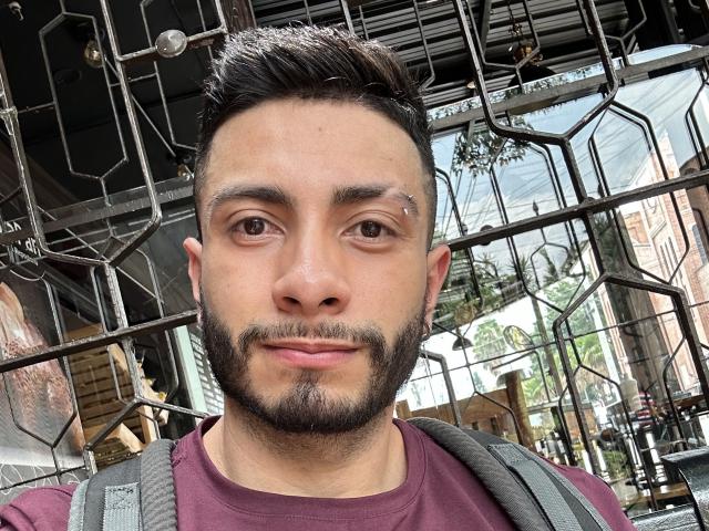 MarkBuffett tem uma barba curta e escura, um pequeno piercing no nariz, e está vestindo uma camisa marrom-avermelhada com uma mochila cinza, olhando para a câmera com um leve sorriso..
