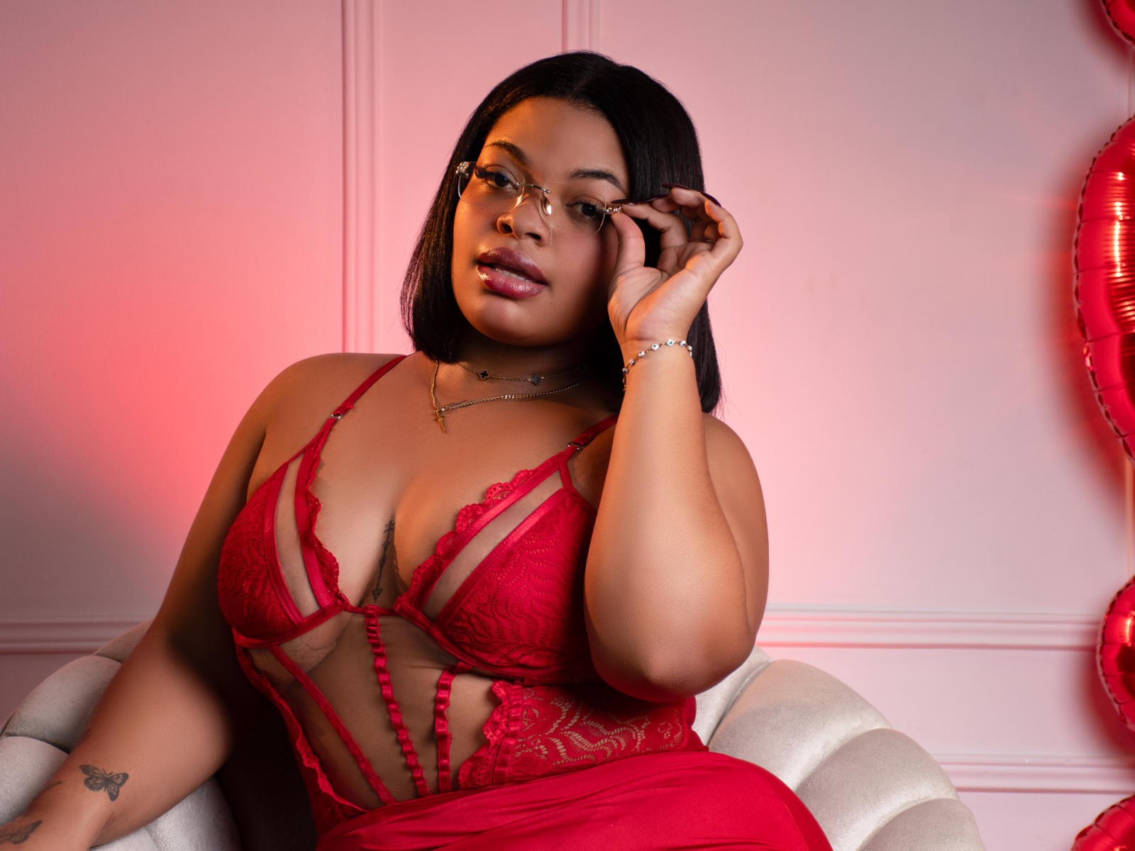 YazminJenner en lingerie rouge en dentelle, montrant ses seins, avec des lunettes, des cheveux noirs, et un éclairage rose-rouge, assise sur une chaise blanche, la main droite touchant son visage..