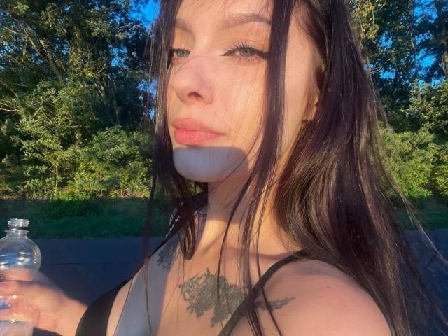 ElisBeautyfull, met lang zwart haar, houdt een waterfles vast, heeft tatoeages op haar borst, draagt een zwarte top, en het zonlicht werpt schaduwen op haar gezicht en borsten..