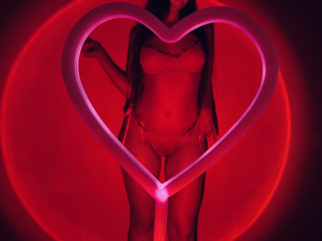 Tisia stands in front of a glowing pink heart-shaped neon frame, wearing red lingerie with a lace bra and thong, highlighting her tits and pussy, with her head out of frame.