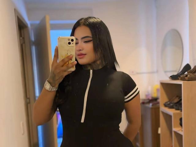 LairaHearvy with long black hair, winking, wearing a black dress with white stripes, holding a phone, in a hallway, her hand on her hip, showing her ass.