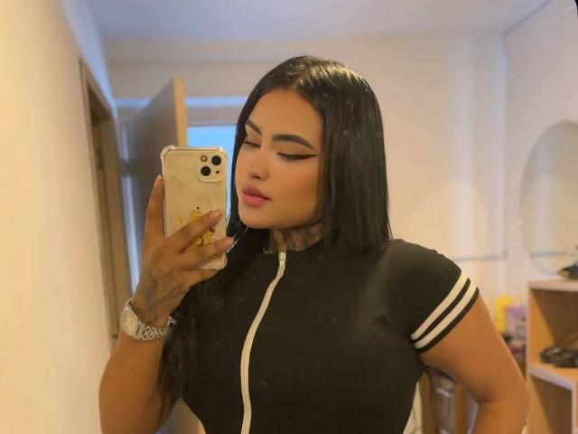 LairaHearvy with long black hair, wearing a black top with white stripes, holding a phone, showing off her tits in a mirror selfie, pink lipstick, tattoo on wrist.