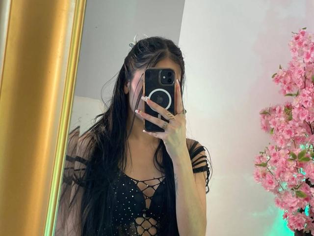 VioletaVelvet with long black hair, lace-up black dress, holding phone covering face, pink cherry blossoms on right, gold mirror on left, taking selfie in mirror.