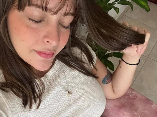 LiaMilan met gesloten ogen, bruin haar, een witte geribbelde top, roze lippen, een kleine neus, een zwarte polsband, een tatoeage op de bovenarm, een roze kussen, een groene plant..