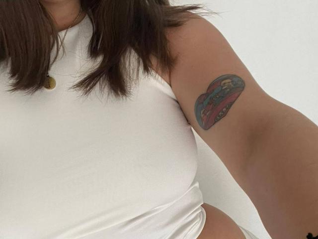 A tatuagem no braço de LiaMilan, um coração colorido com um carro vermelho, é visível acima de sua blusa branca sem mangas. Seu cabelo castanho está solto, e ela usa um colar de ouro..