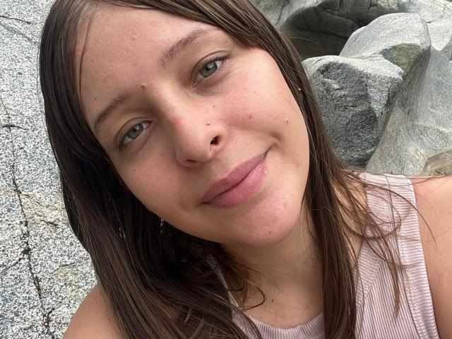 Il viso di LiaMilan, con i capelli castani, gli occhi verdi, le labbra rosa, un piccolo piercing al naso e un leggero sorriso. Indossa una canotta a righe rosa, con delle rocce sullo sfondo..