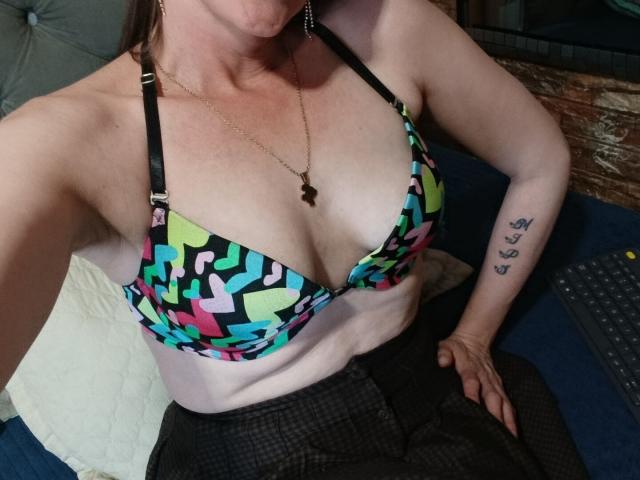RubiAnderson with pale skin, wearing a colorful geometric patterned bra, exposing her tits, black checkered pants, tattoo on right arm, gold necklace, and earrings, sitting on a blue couch.