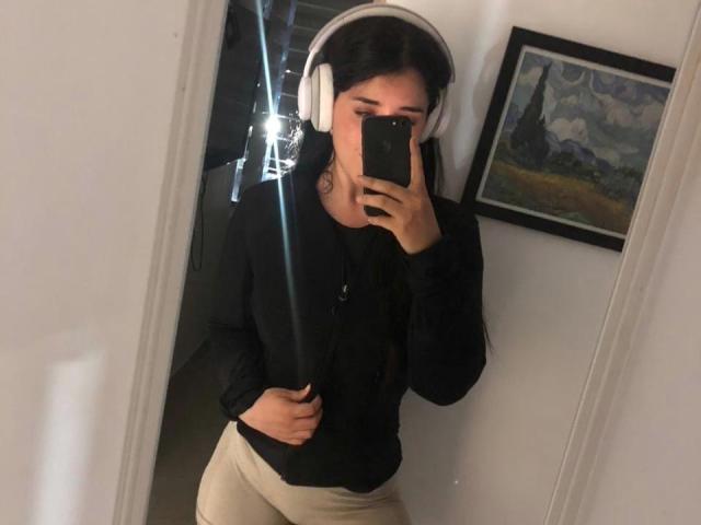 VickyMegan wearing white headphones, black long-sleeve shirt, and beige pants, taking a mirror selfie with her phone, black hair, medium build, and a slight smile.