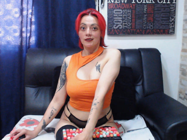 SamanthaSwan with red hair, orange halter top, black panties, tattoos, and black thigh socks, sitting on a black leather couch, holding a bowl of red and white candies.