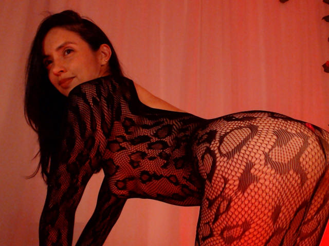 BelladonnaLips with long dark hair, wearing a black lace fishnet bodysuit, bent over, showing her ass and looking back with a slight smile, in red lighting.