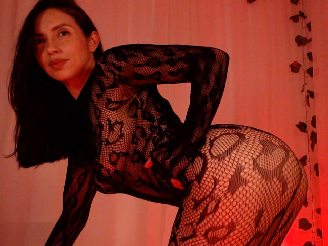 BelladonnaLips with dark hair, wearing a black lace bodysuit, bending forward, showing her ass through the patterned mesh, red light on her.
