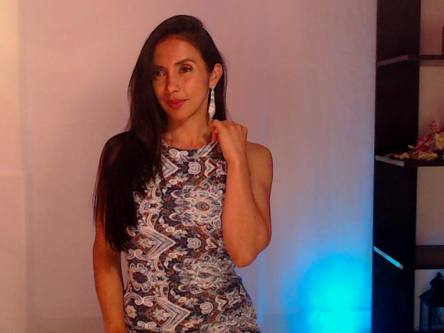 Belladonna: Eine Frau mit langen, dunklen Haaren, die ein blau-weiß gemustertes Kleid trägt, eine silberne Ohrringe hält und mit einem leichten Lächeln posiert. Auf dem Foto ist ihr Hintern zu sehen..