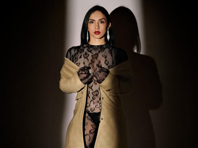 BelladonnaLips stands in shadow, wearing a black lace bodysuit, beige coat off-shoulder, showing her tits through the lace, red lipstick, black hair, and silver earrings.