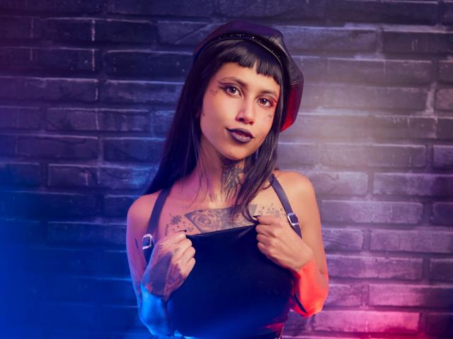 NyxVega with black hair, wearing a red cap, black tank top, and suspenders, holding a tablet, tattoos on chest and neck, dark lipstick, blue and pink neon light.